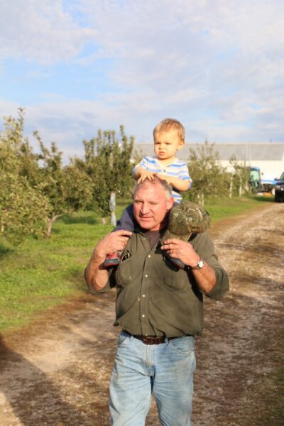 A man carrying a toddler on his shoulders while walking through an orchard, surrounded by trees and a rural landscape under a partly cloudy sky. The man is dressed in a green shirt and blue jeans, with a camouflage cap, while the child wears a striped shirt.