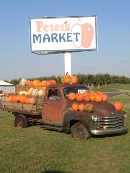 Rustic pickup truck loaded with pumpkins and gourds parked in front of Peter's Market sign, set against a clear blue sky. Ideal for fall harvest promotions and local farm market imagery.