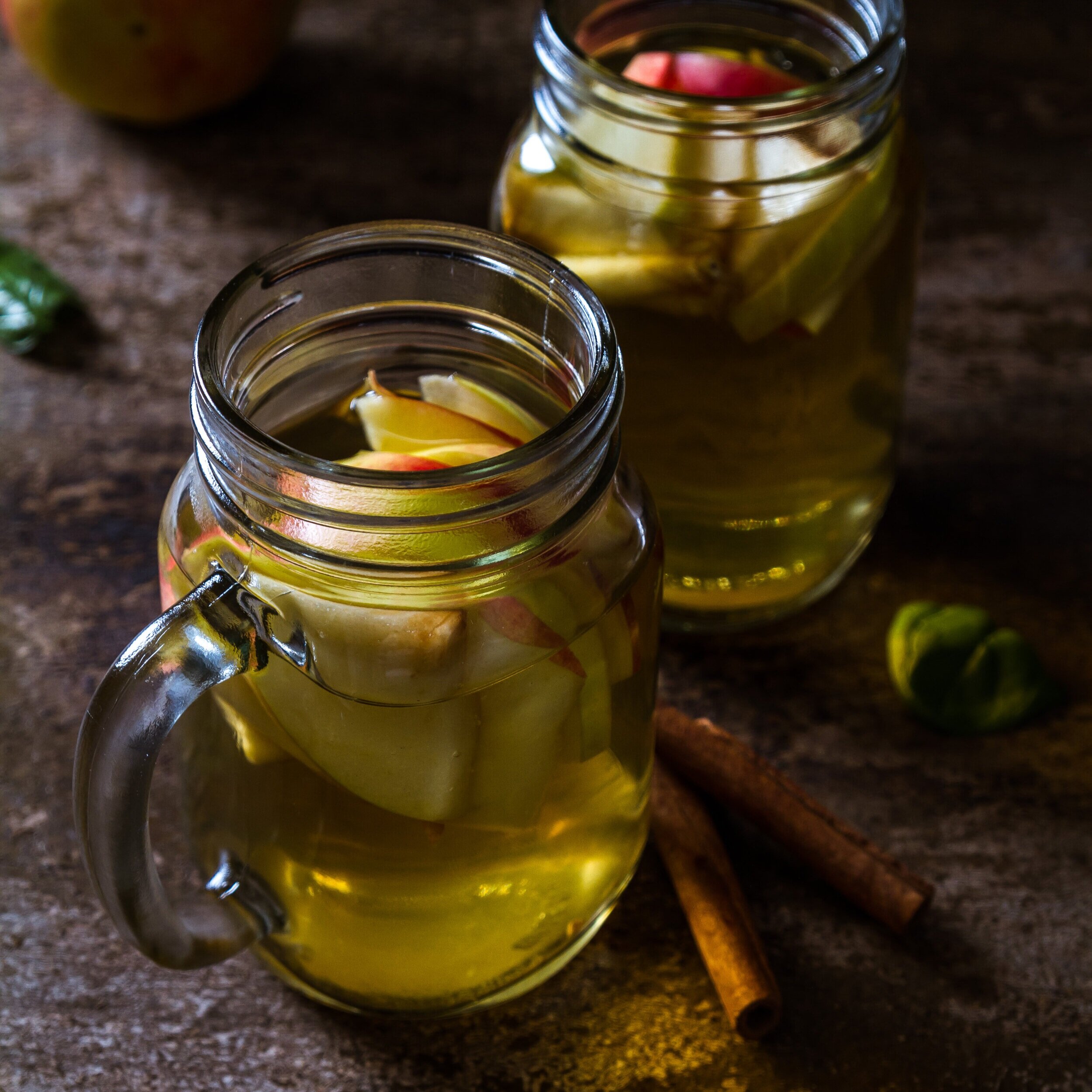 Two glass jars filled with apple slices immersed in a warm, golden beverage, surrounded by cinnamon sticks and green leaves on a rustic wooden surface. Perfect for a cozy fall drink or a refreshing apple-infused beverage.