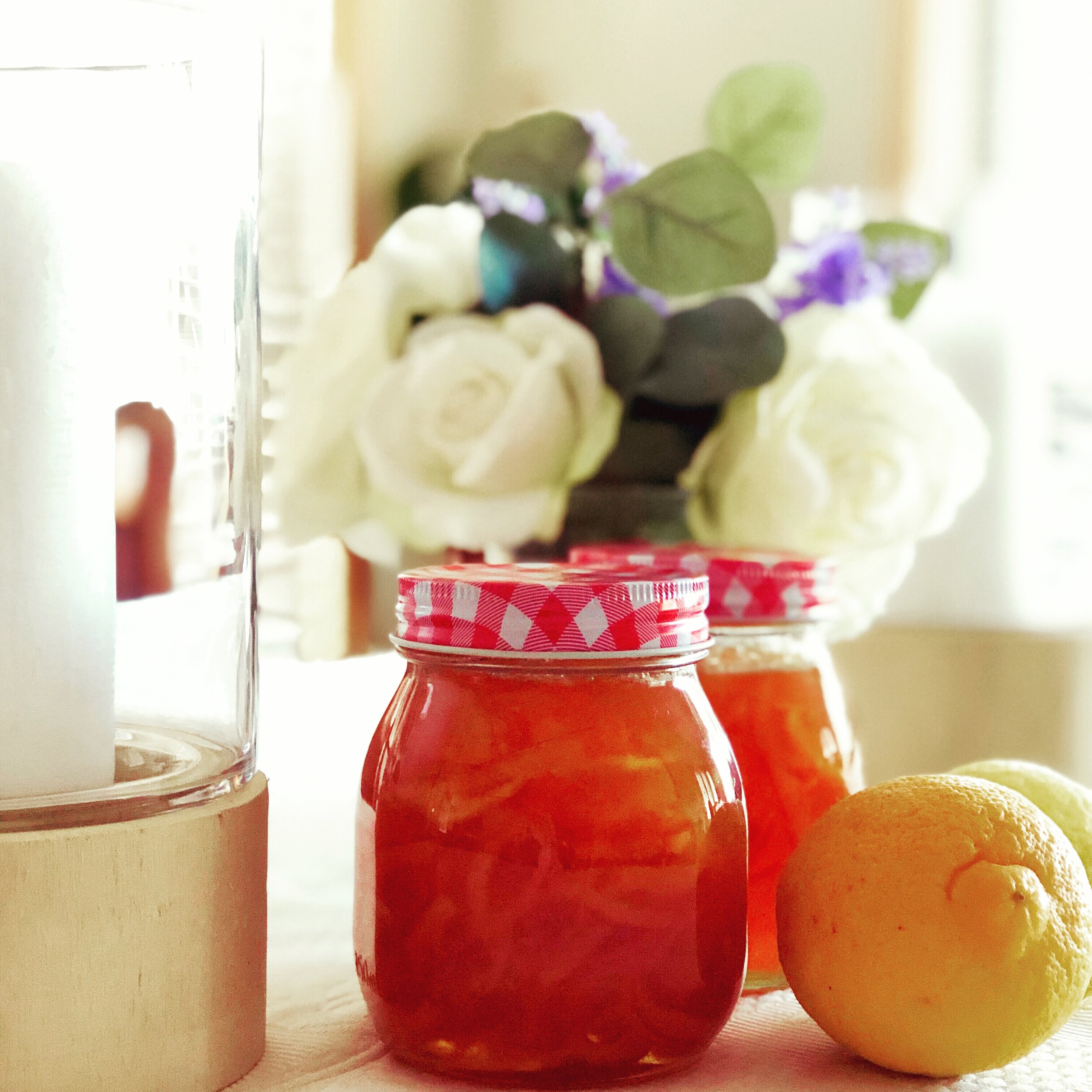 Jars of homemade citrus marmalade with red gingham lids, placed on a table next to fresh lemons and a decorative floral arrangement, creating a cozy kitchen atmosphere.