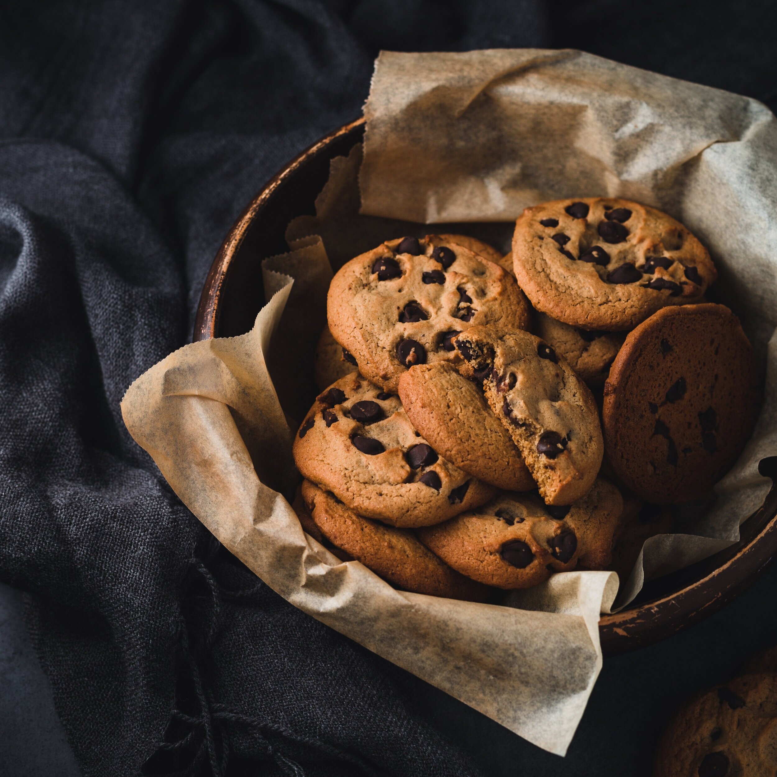 Freshly baked chocolate chip cookies arranged in a wooden bowl lined with parchment paper, set against a dark, textured background.